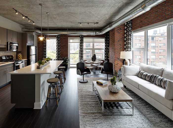 deco industrielle dans in salon avec canapé blanc, table basse em bois et metal, plafond effet beton, mur en briques, tuyauterie avec cuisine adjacente