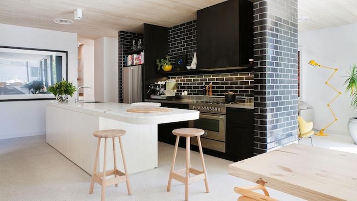 cuisine moderne blanche et noire, meuble cuisine et mur en briques noir, ilot central blanc avec des tabourets en bois, sol gris