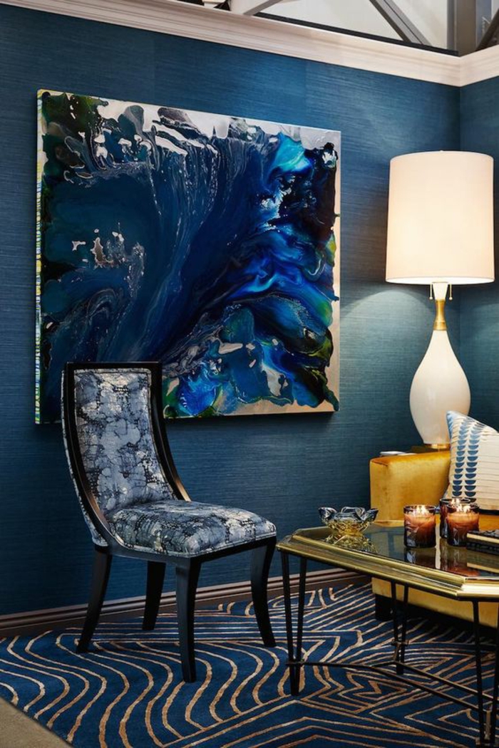 amenagement salon avec chaise fauteuil en bleu et blanc, grand panneau décoratif en nuances du bleu et du noir, table carrée avec plan doré et des pieds en métal noir, tapis aux motifs dunes en bleu marin et beige, murs en bleu canard 