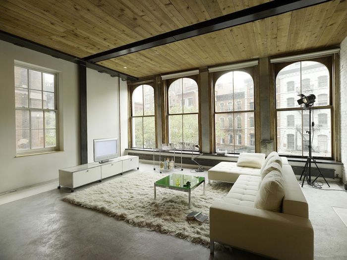 exemple de déco loft industriel dans un salon spacieux avec canapé beige, tapis moelleux blanc, table basse en metal et verre, meuble tv blanc, grande baies vitrées, plafond en bois