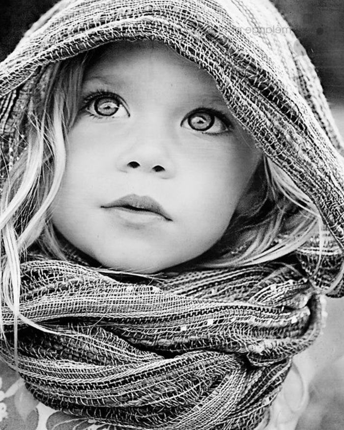 photo en noir et blanc, enfant fillette aux cheveux blonds, avec la tete couverte d'un châle en lin coloré, grands yeux innocents et questionnant 