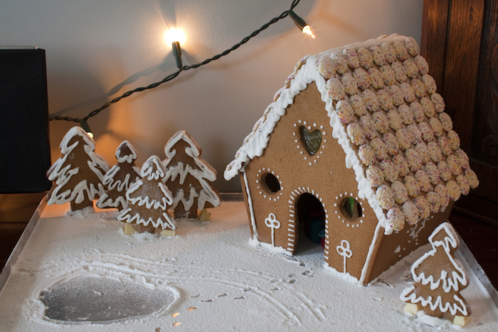 une maison en pain d épices avec décoration de glacage blanc et petits bonbons au chocolat blanc décorés de billes colorés, biscuits en forme de sapin, sucre glace en guise de neige