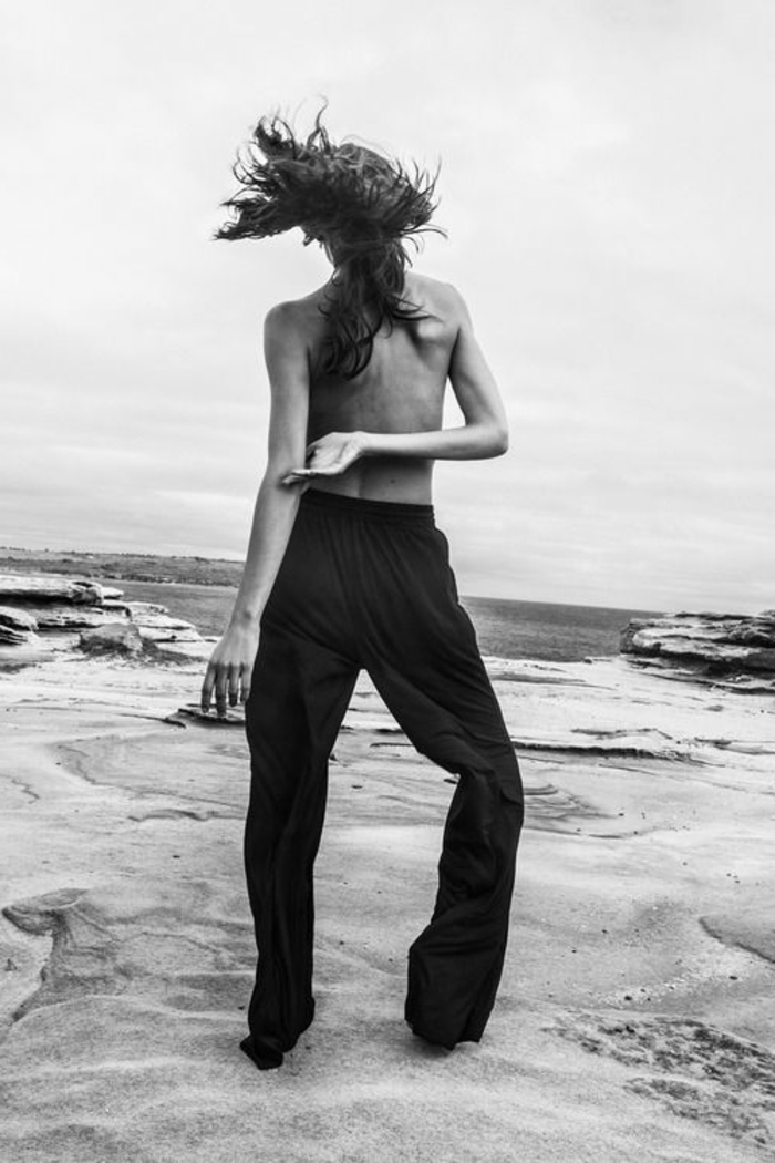 photo femme noir et blanc, dame avec pantalon noir, buste nu, en face de l'océan, cheveux longs qui volent au vent, paysage naturel inspirant