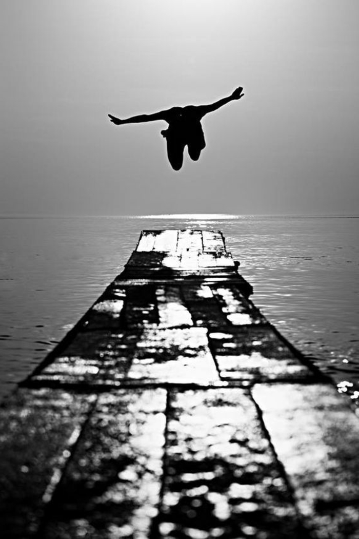 photo femme noir et blanc, homme qui du quai saute dans la mer, bras grands ouverts comme pour un vol dans le vide 