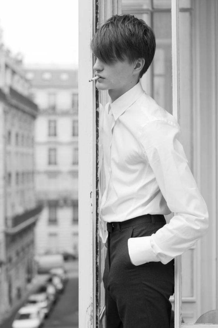 photos noir et blanc, portrait noir et blanc, jeune homme avec la figure de mannequin qui fume sur une terrasse parisienne, coiffure de rebelle, scrute l'édifice en face 