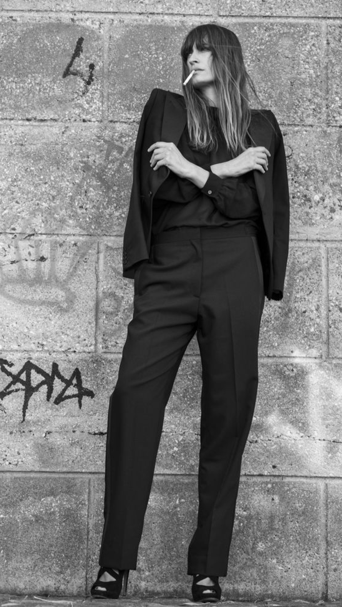 image noir et blanc, portrait en noir et blanc, femme en tenue habillée veste avec pantalon noir, qui fume une cigarette devant un mur avec des graffitis 