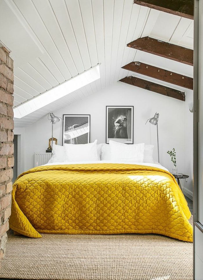 mur en briques rouges, lit avec parure jaune, poutres en bois, plafond et murs blancs, comble aménagé