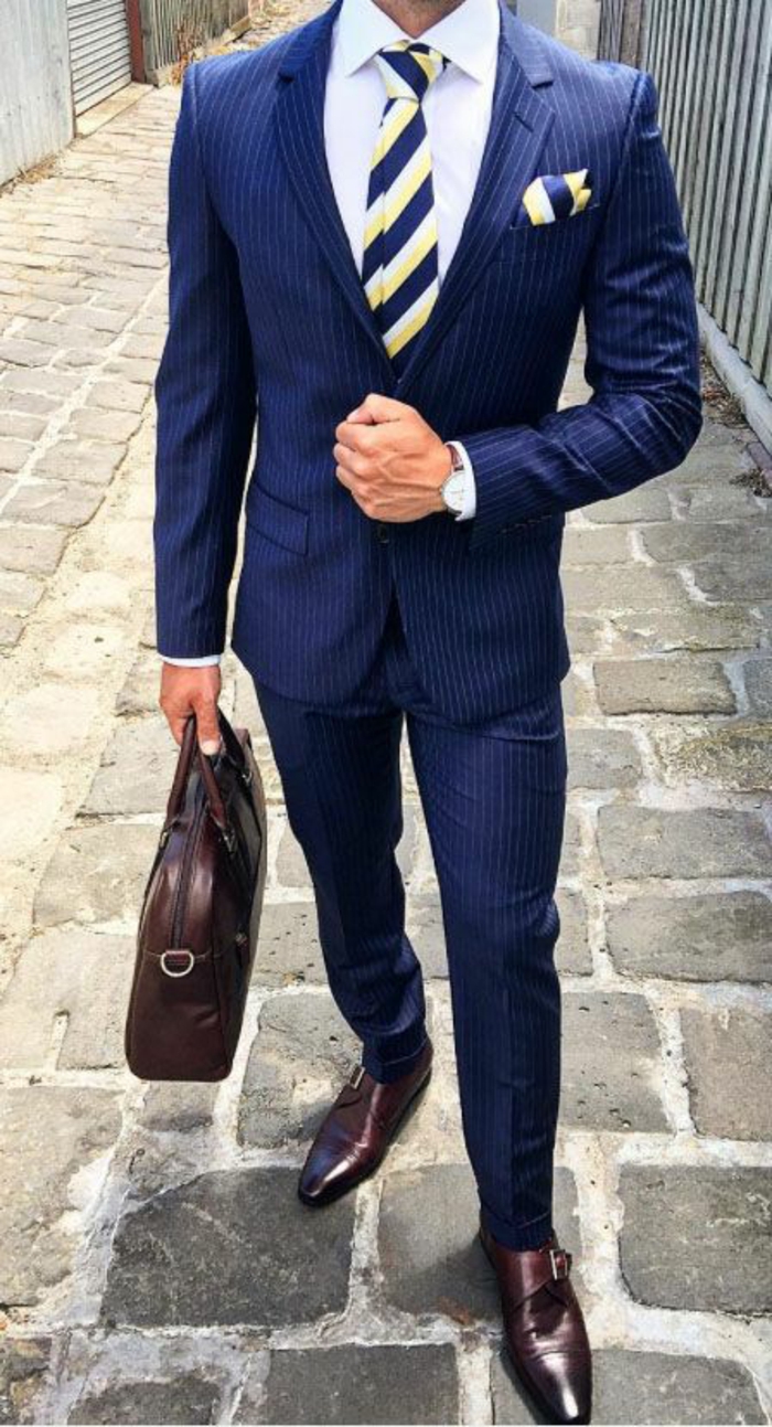 homme d'affaires, costard bleu avec cravate aux rayures diagonales en bleu roi et blanc, chaussures en couleur bordeaux et porte-sac élégant en couleur bordeaux