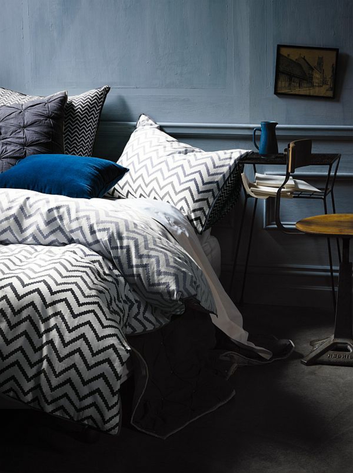 coussin bleu, linge de lit à zigzags, mur couleur colombe, petite table haute, chaise en bois et fer industrielle