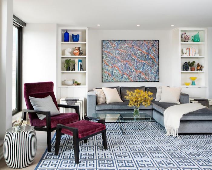 etageres blanches, canapé gris d angle, tapis à motifs géométriques, table basse en verre, chaise et tabouret couleur framboise, tableau cadre mural