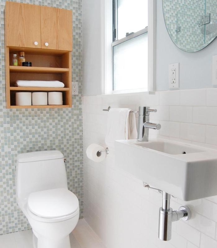 agencement salle de bain avec un mur en carrelage gris et blanc et reste du carrelage blanc, lavabo et wc blanc, miroir rond, meuble au dessus de toilette