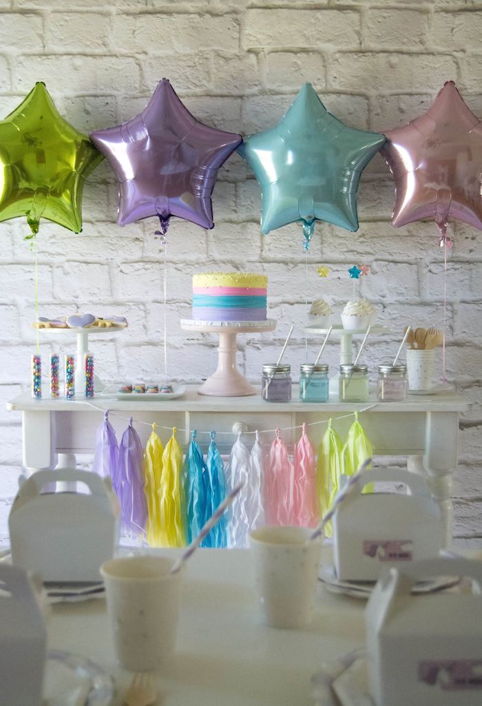 anniversaire-licorne-gâteau-arc-en-ciel-guirlande-à-glands-ballons-aluminium-étoiles-buffet-mur-en-brique-blanche un candy bar en tons pastel décoré de ballons aluminium en forme d'étoiles, une guirlande de tassels et un gateau licorne au glaçage arc-en-ciel