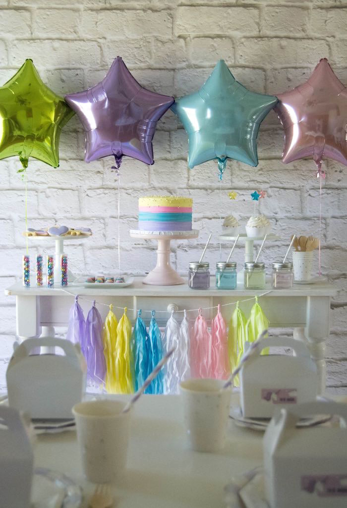 un candy bar en tons pastel décoré de ballons aluminium en forme d'étoiles, une guirlande de tassels et un gateau licorne au glaçage arc-en-ciel