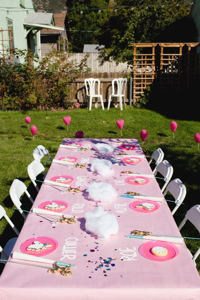idées déco pour un anniversaire en plein air, jolie décoration de table avec une nappe rose personnalisée avec les noms des invités inscrit dessus et un accessoire licorne près de chaque assiette