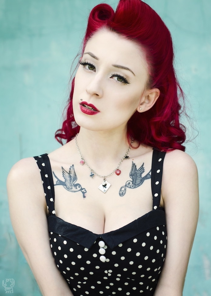 modele de coiffure rockabilly pin up sur cheveux framboise avec des boucles et banane rockabilly en haut, tatouage oiseau et robe noire à pois blancs