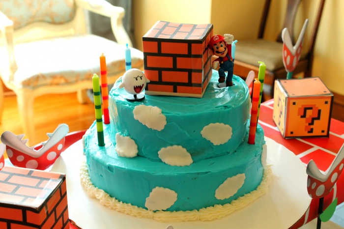 un gateau d'anniversaire à deux étages sur thème super mario recouvert de glaçage bleu décoré de nuages en pâte à sucre et de figurines super mario