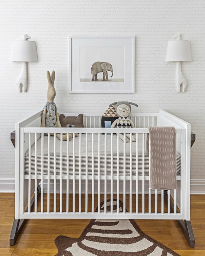 lit bébé blanc en bois, tapis en forme de peau de zèbre, deux luminaires appliques avec des abat-jours blancs en forme de girafes, parquet en jaune, tableau avec éléphant gris au cadre blanc 