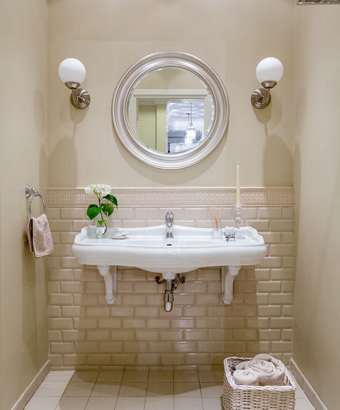 lavabo blanc vintage dans une salle de bain petite surface beige, miroir rond, panier rangement et lampes murales