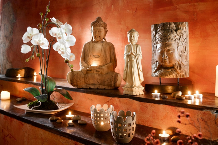 couleur zen, bouddha zen, deco salon zen, mur en orange, étagères en marron avec plein d'objets déco, statuette de Bouddha, bougeoirs en céramique et en métal, orchidée blanche dans un pot beige en style zen 