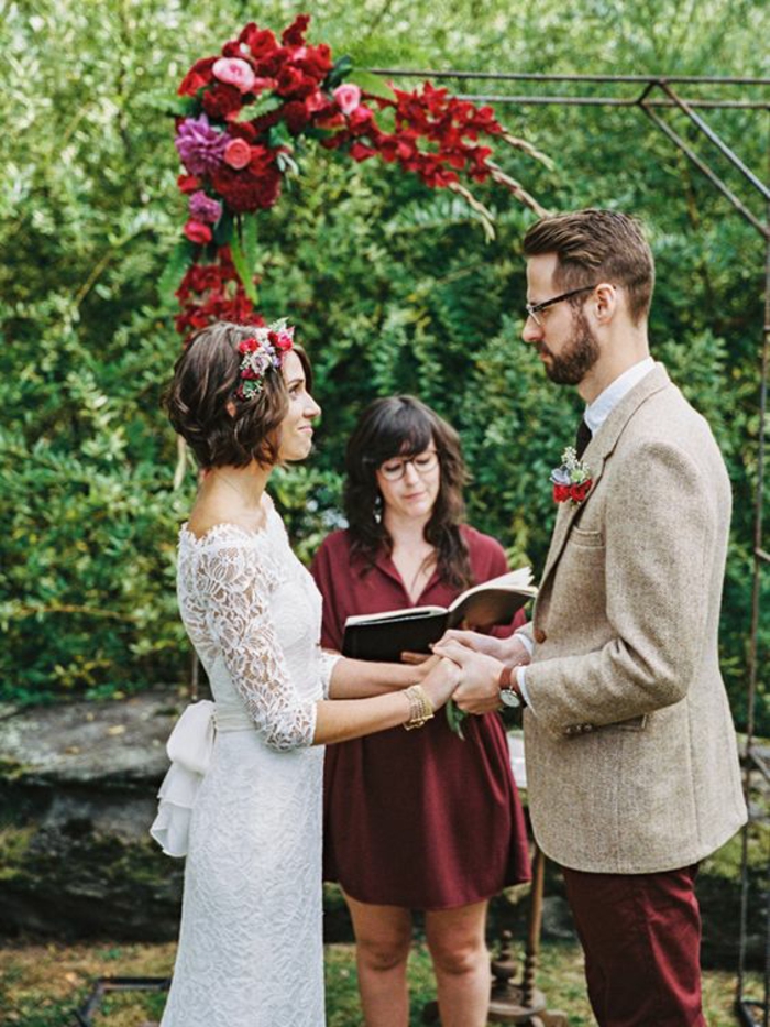 des mariés qui se disent oui devant une arche avec des roses rouges et roses, coiffure mariage cheveux courts, diadème mariée en fleurs et feuilles