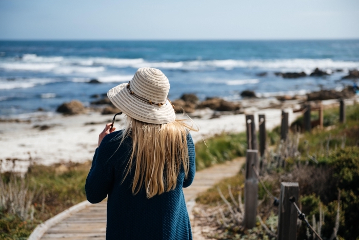 jeune femme en promenade au bord de l'océan habillée en gilet en crochet bleu foncé avec accessoires tendance capeline et lunettes de soleil