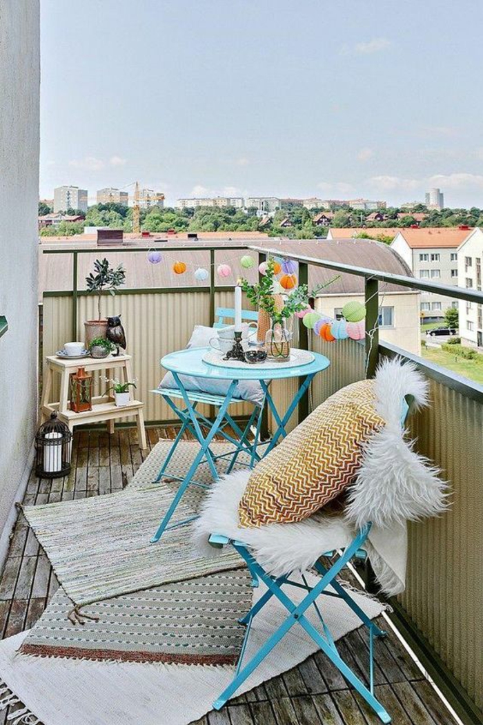 idée déco appartement, balcon décoré avec guirlande lumineuse avc boules colorées, trois tapis au sol de taille diverse en réséda et ivoire, deux chaises en métal bleu pastel pliables, table ronde et haute en métal bleu pastel