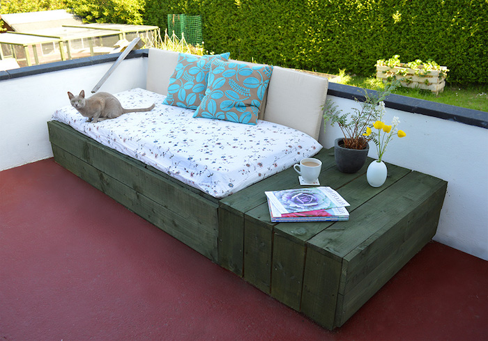 canapé en palette de bois avec des planches vert olive, coussin d assise blanc et coussins décoratifs gris et bleu, pots de fleurs