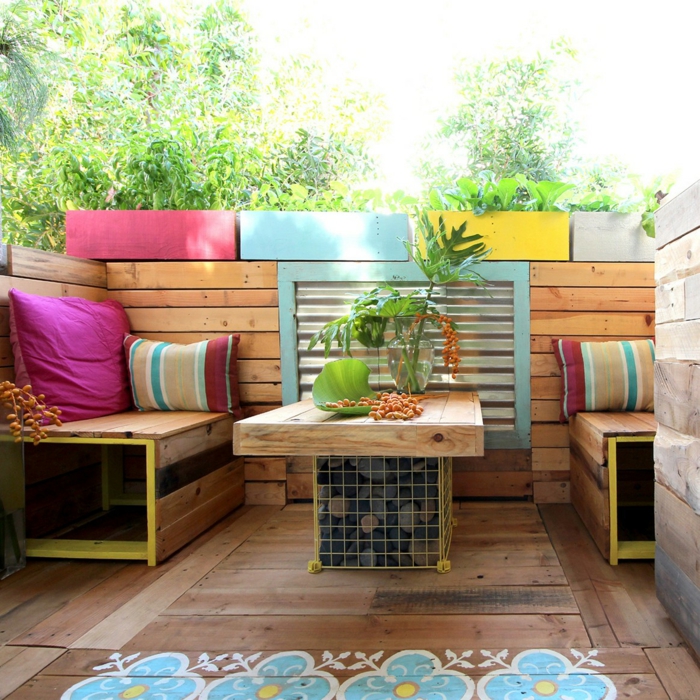 zone de terrasse avec des meubles de jardin en palettes, table et canapé palette, quatre pots rectangulaires avec des plantes en fuchsia, bleu pastel, jaune et couleur ivoire 