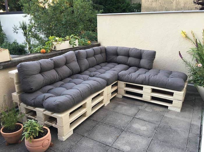 meuble avec palette en bois avec coussin d assise gris sur une terrasse couverte de dalles de béton, pots de fleurs