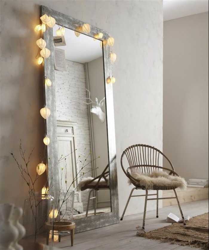 déco murale avec un grand miroir au cadre couleur argent, orné d'une guirlande lumineuse de lanternes boules blanches, style de pièce minimaliste, fauteuil en métal au dossier rond, tapis couleurs terre 