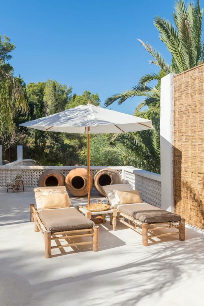 deux chaises-longues en bois beige recouverts de matelas en marron et doré, parasol tissu blanc et bois, idée déco terrasse en style méridional avec des palmiers