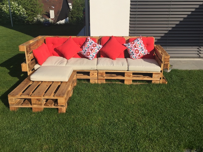 meuble avec palette en bois, un canapé bois brut avec des coussins d assise beige et coussins décoratifs rouges, gazoon vert