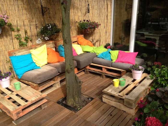 terrasse en bois avec un canapé bois de palette avec coussins d assise gris et coussins décoratifs colorés, petites tables de service en palettes, brise vue en bambou