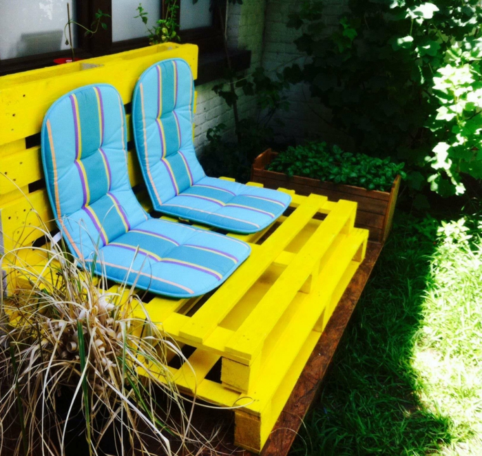 banquette en palette peinte en jaune flashy et recouverte de deux matelas en bleu pastel et en jaune, meubles de jardin en palettes posés a l'ombre d'un arbre 