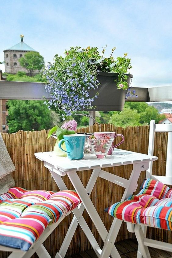 balcon fleuri, idée déco terrasse, chaises et table pliable en bois blanc PVC, garde-corps recouvert de bambou pour préserver l'intimité, coussins meubles aux rayures verticales en couleurs vitaminées 