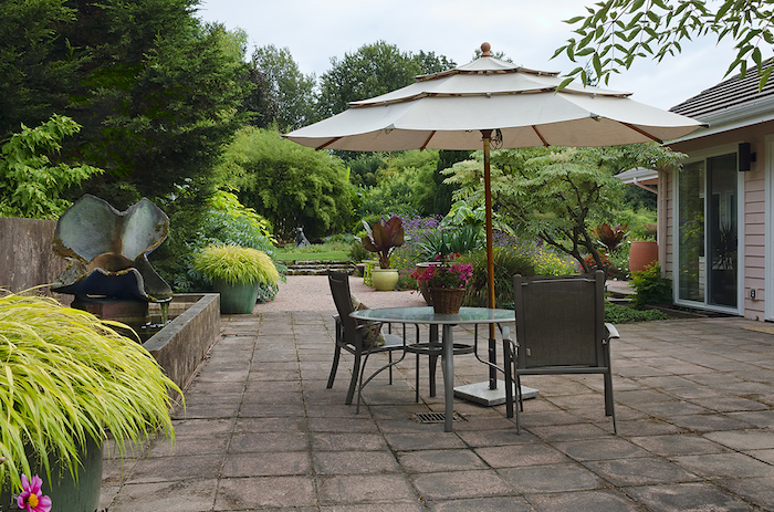 pavage exterieur avec une table ronde et chaises, basssin d eau simple, plusieurs arbustes, arbres et plantes en pots et bacs