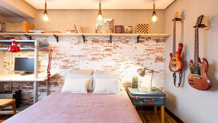 mur de briques avec une étagère bois, linge de lit blanc et rose, bureau style industriel et guitares décoratives, table de nuit en malle recyclée