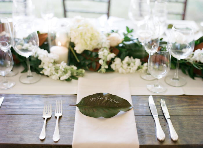 feuille verte décorée de nom invité noté au feutre blanc, chemin de table et serviette blanche et table bois brut, centre feuillage et fleurs blanches