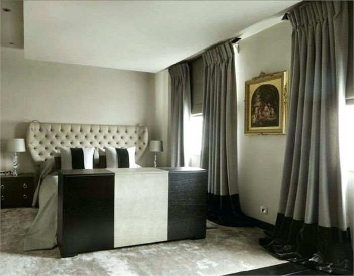 une chambre en style imposant, lit avec tête de lit baroque, rideaux gris, peinture à l'encadrement doré et tapis gris