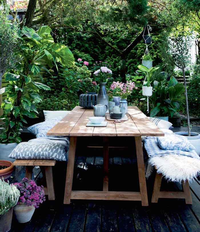 bancs bois et table en bois brut sur une terrasse bois brut, jardin envahi par une végétation verte, bouquets de fleurs champetre, plusieurs coussins et plaids