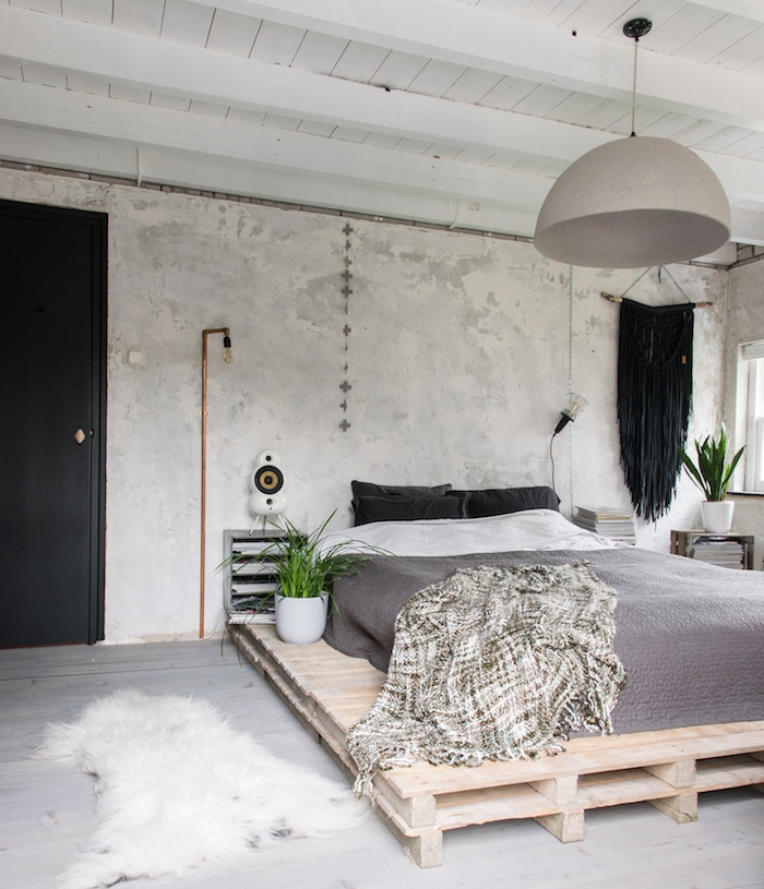 plafond ossature bois apparente, lit en palette avec matelas et linge de lit gris et blanc, mur béton, parquet gris, suspension grise, plante verte
