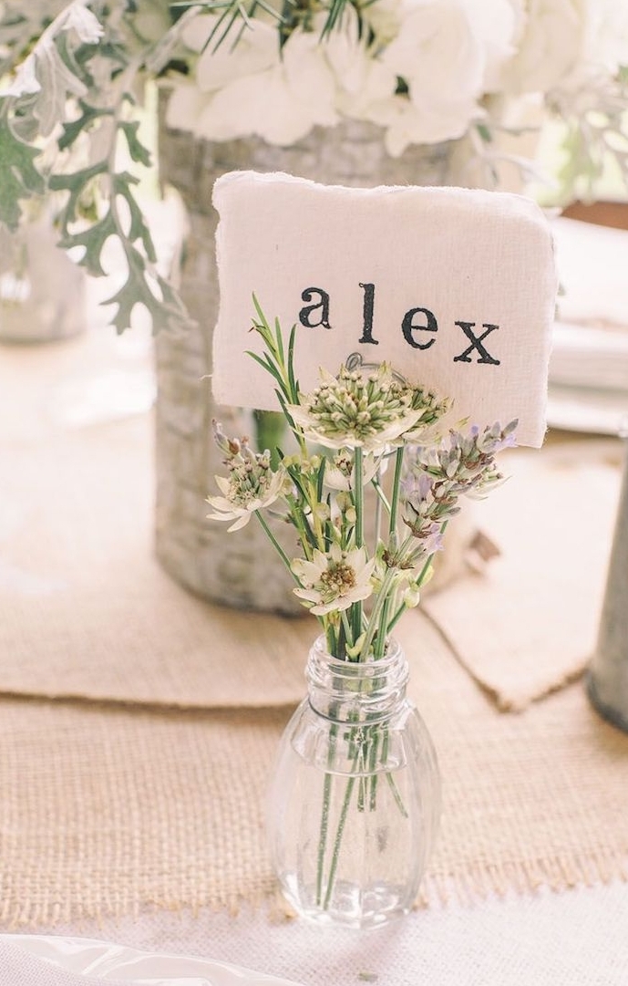 petite bouteille en verre avec un petit bouquet de fleurs à l intérieur avec étiquette prénom invité, chemin de table en jute, centre floral