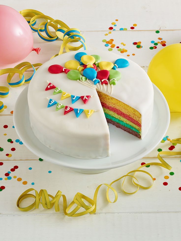 joli gâteau arc en ciel recouvert de fondant blanc et décoré avec des ballons et des guirlandes modelés en pâte à sucre