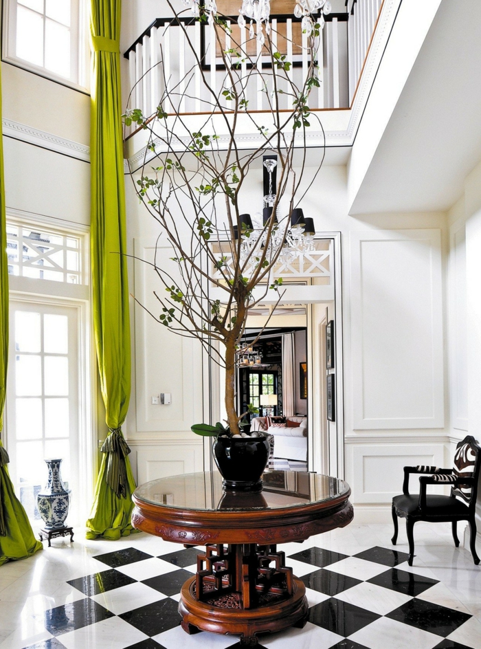 entrée blanche, grande fenêtre et rideaux verts, petite table ronde avec un arbre décoratif, carrelage damier, chaise baroque
