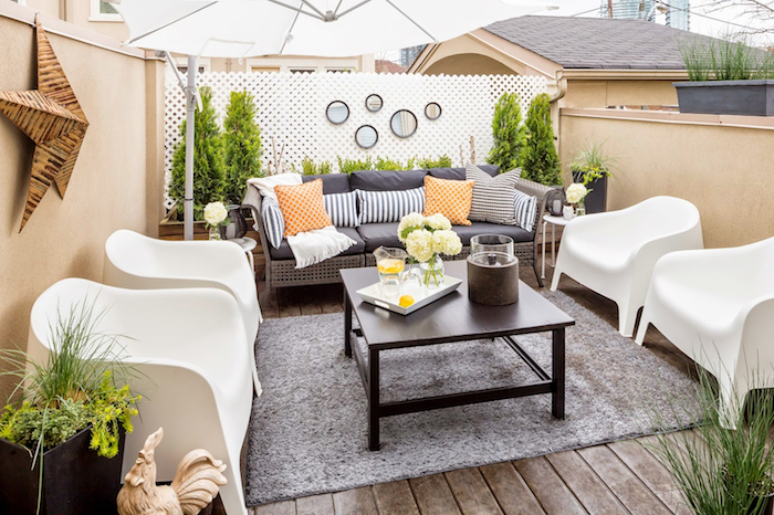 amenagement patio sur une terrasse exterieur avec canapé décoré de coussins, chaises blanches, tapis gris sur un sol en bois, quelques plantes en pots