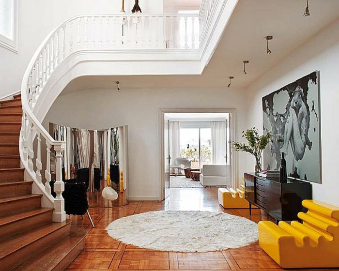 tapis rond, sol marron, escalier tournant, fauteuils jaunes, commode noire, peinture artistique géante