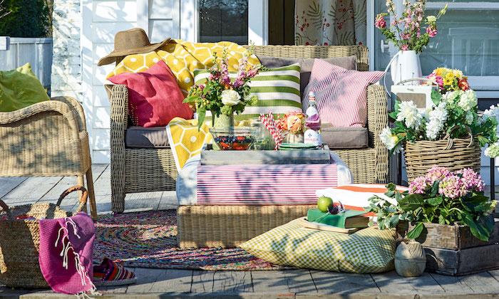 amenagement petite terrasse exterieur en bois naturel avec canapé et chaise rotin, coussins colorés et tapis tressé coloré, plusieurs bouquets de fleurs