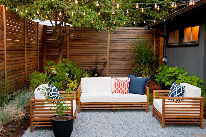 aménagement cour extérieur avec salon de jardin bois composé de fauteuils et canapé bois, revêtement sol en gravier, clôture bois composite, guirlande lumineuse