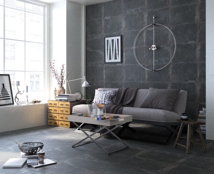 salon de style industriel avec mur gris foncé et grande fenêtre, canapé gris couvert de coussins et plaid décoratifs en nuances de gris