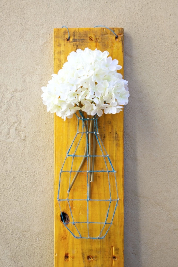 joli vase décorative en fil bleu et clous remplie de fleurs blanches sur une planche de bois peinte en jaune